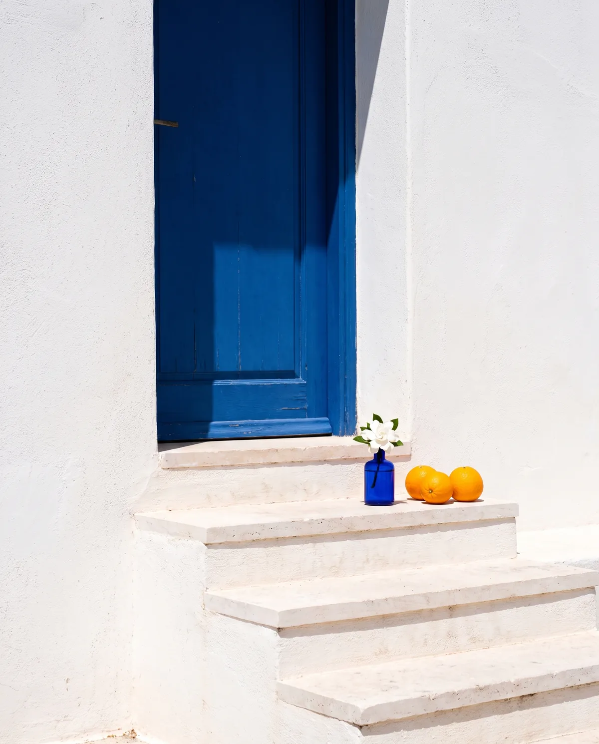 Oranges and a small cobalt glass vase with a white flower on white stone steps in hard sunlight with blue shadows